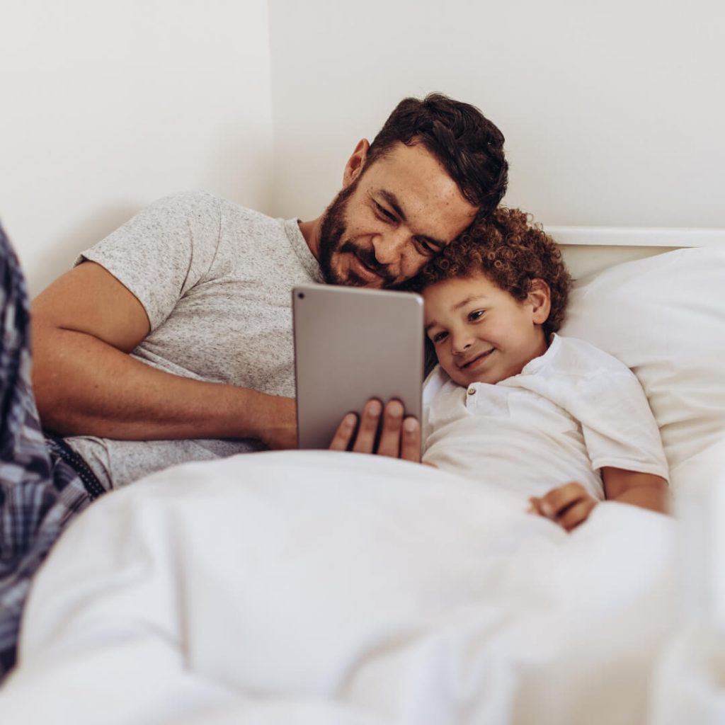 dad snuggling son in bed with tablet