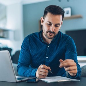 man looking at papers in clipboard with open computer