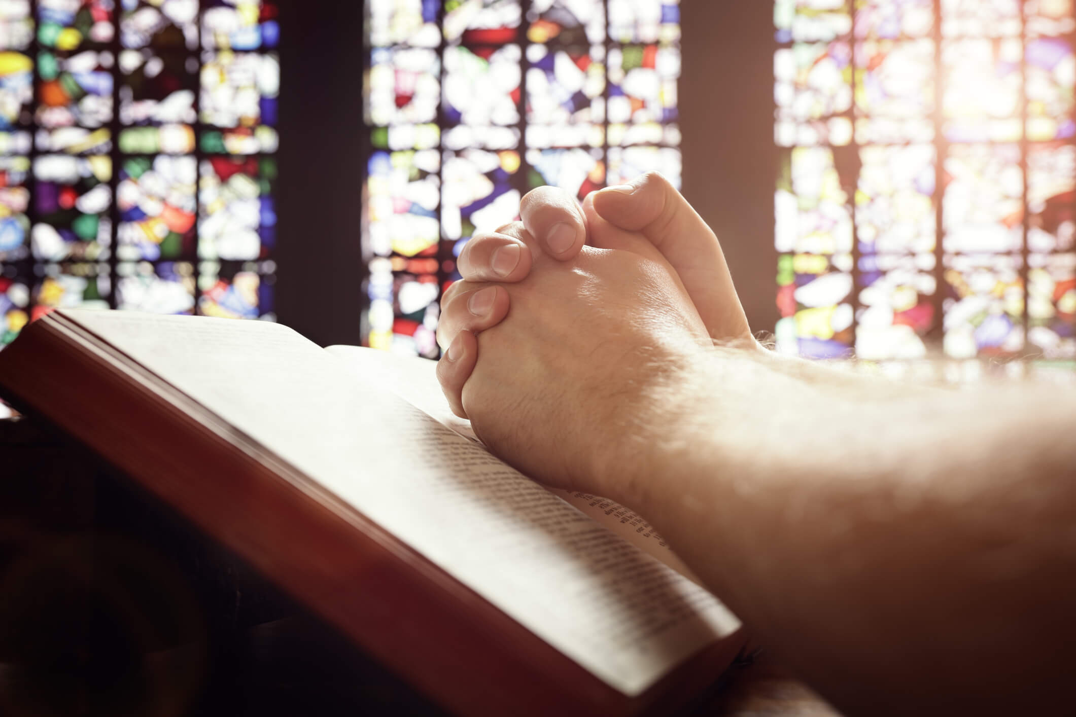 Folded hands of a man praying in front of stained glass.