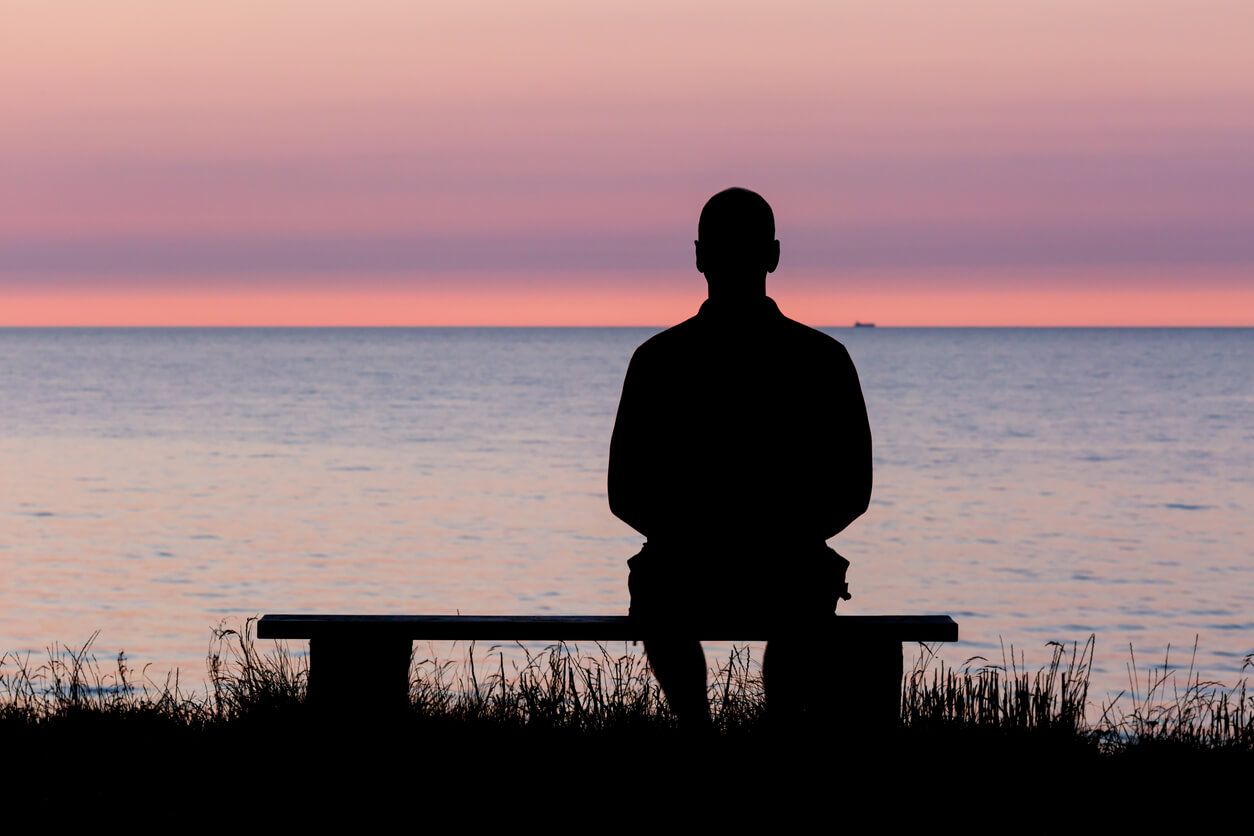 Silhouette of a man sitting on bench overlooking a lake at sunset.
