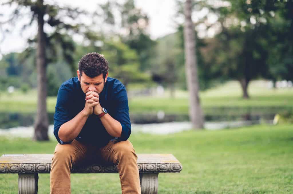 A man praying outside on a park bench.