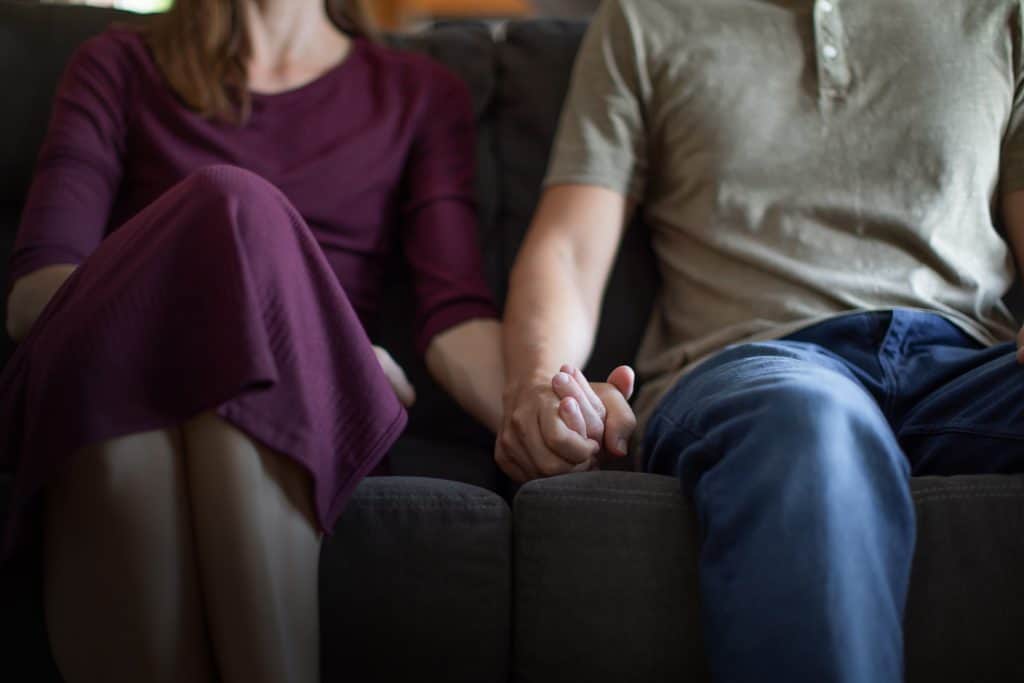 A husband and wife sit on the couch holding hands.