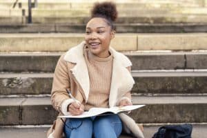 Female student writing in a notebook, making a plan on stairs in city.