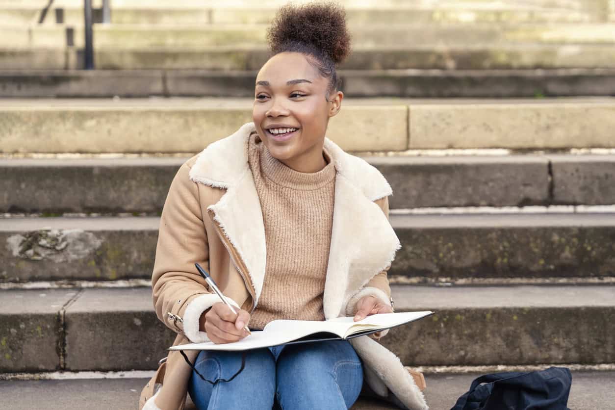 Female student writing in a notebook, making a plan on stairs in city.