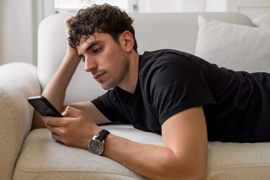 Young male lying down on sofa and surfing on net with his smart phone