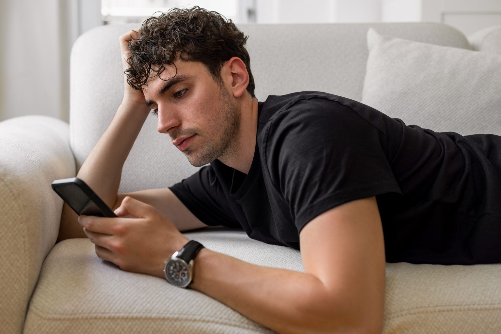 Young male lying down on sofa and surfing on net with his smart phone